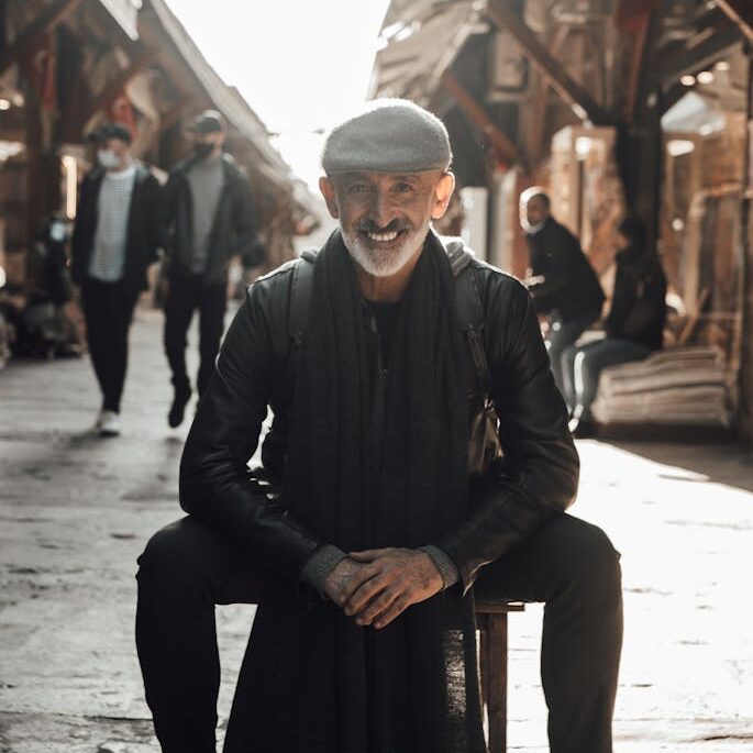 Ethnic happy middle aged male in casual clothes and hat sitting on stool on street market in town in sunny day and looking at camera